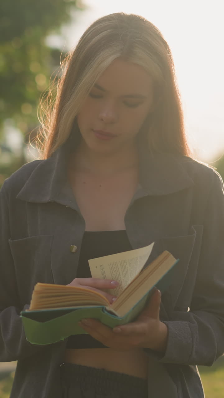 dama con ropa gris de pie al aire libre hojeando las páginas de un libro, mientras la luz del sol ilumina suavemente su figura y el fondo presenta árboles borrosos y un edificio