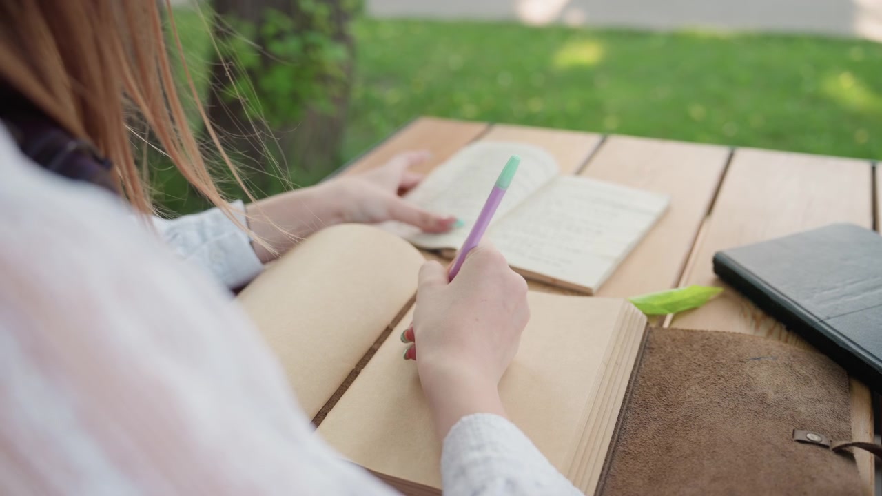 Close up of young woman writing in open notebook with her left hand while holding another book steady on wooden table. purple pen is held between pages and various notebooks including one
