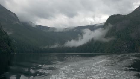 A-misty-Alaskan-fjord-reveals-a-mountain-range-sitting-on-the-water-beyond-the-wake-of-a-ship