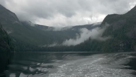 a misty alaskan fjord reveals a mountain range sitting on the water beyond the wake of a ship