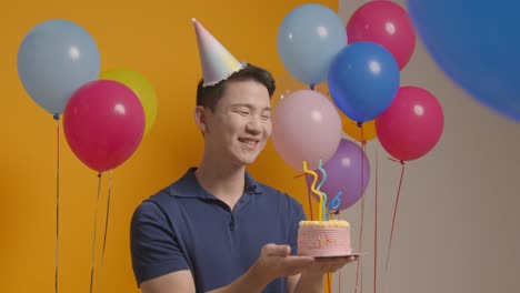 studio portrait of man wearing party hat celebrating birthday blowing out candles on cake