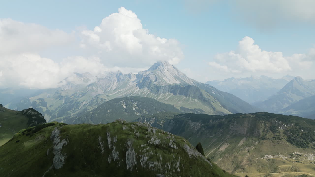 Drone aerial view over a small grass and rocky hill until a large mountain peak stretches out in the background, still lit by the sun. The blue sky is visible behind the clouds.