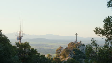 a wide shot of a cross on a hill in front of the monastery of san salvador, mallorca, framed by trees in the foreground