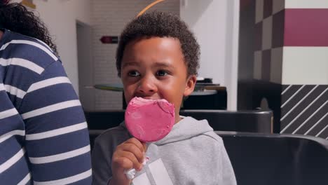 close-up view of a young biracial black boy eating a pink strawberry-flavored artisan ice cream indoor next to his mother, wearing a gray hoodie