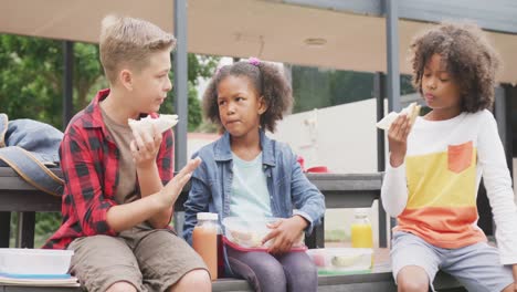 video of three diverse schoolchildren eating packed lunches, talking in schoolyard