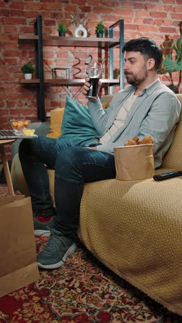 man relaxing on a couch, drinking soda and eating fried chicken