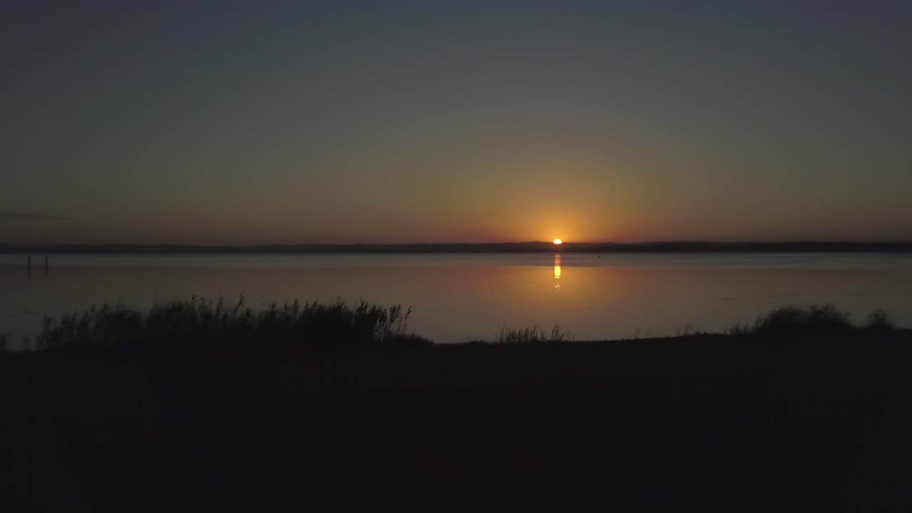 Spectacular sunset over a large calm lake with vegetation as silhouette in the foreground - downward forward aerial motion switching to sideview panning of silhouetted landscape and setting sun.