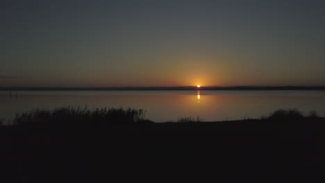 spectacular sunset over a large calm lake with vegetation as silhouette in the foreground - downward forward aerial motion switching to sideview panning of silhouetted landscape and setting sun.