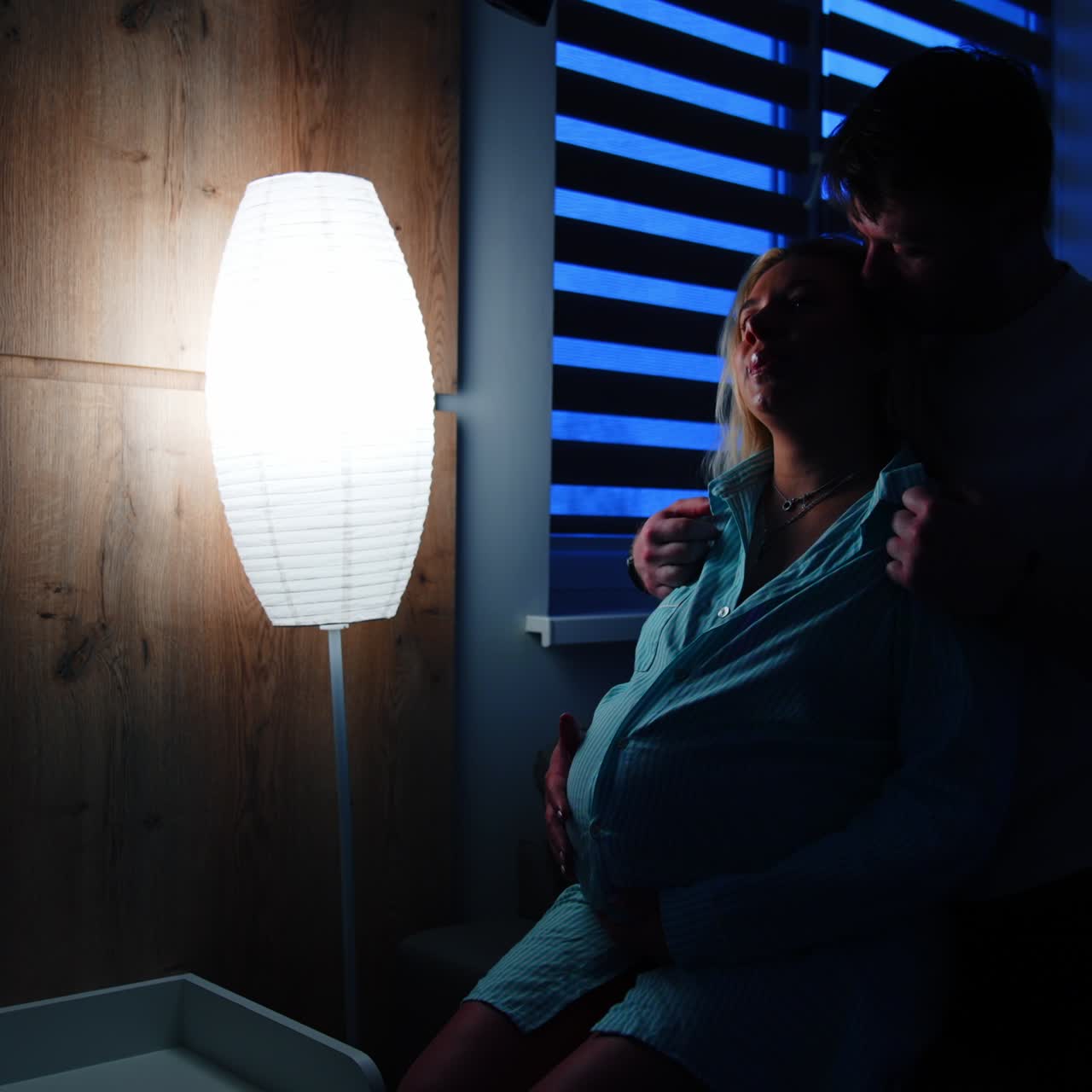 Husband caressing his pregnant wife sitting on the bed. Woman prepares for childbirth in hospital. Dark room backdrop