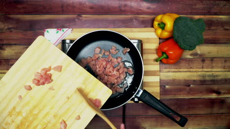 una vista superior de agregar el pollo al aceite caliente en la sartén, colocado en una estufa, dos grandes pimientos amarillos y rojos y un brócoli verde sobre la mesa