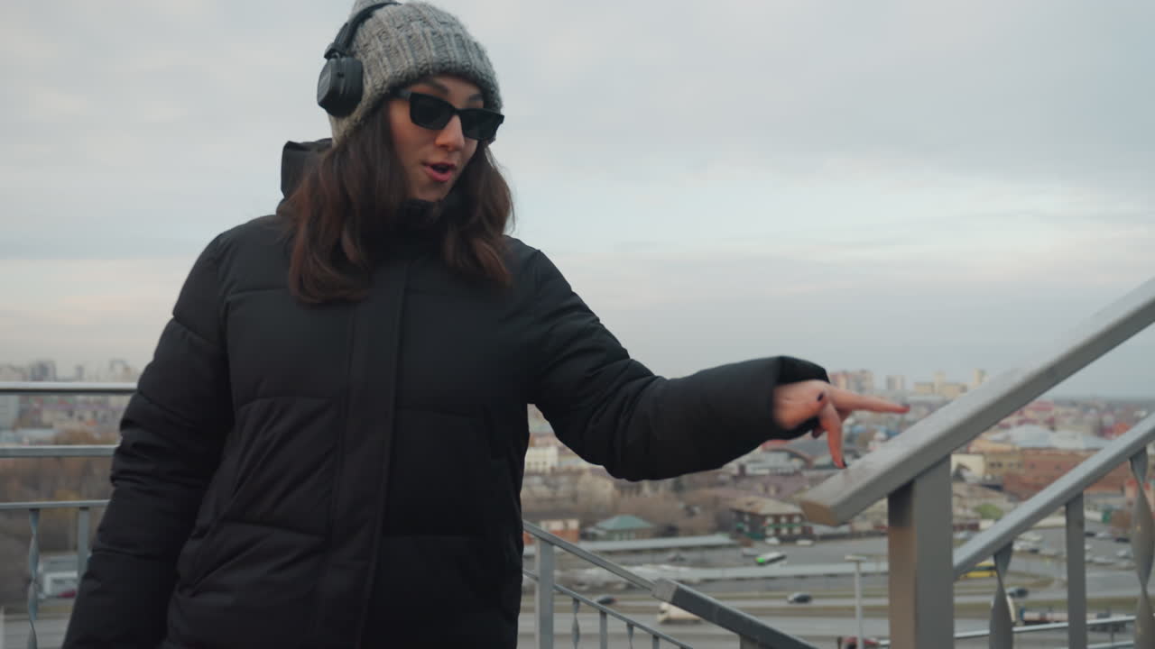 Staircase motion woman dancing with sunglasses and headphones, playful hand gestures while holding railing, pointing toward skyline, cloudy sky bokeh, energetic casual steps, commuter vibe on rooftop