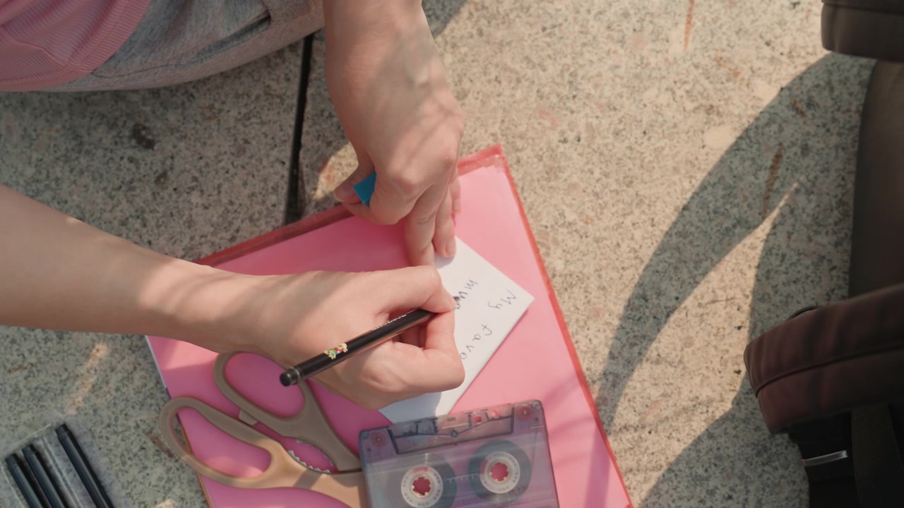 Giovane ragazza scrive su un biglietto con la scritta "La mia musica preferita" visibile, usando vari articoli di cancelleria come forbici e penne. La scena è ambientata all'aperto durante il giorno, con un focus ravvicinato sulle sue mani.