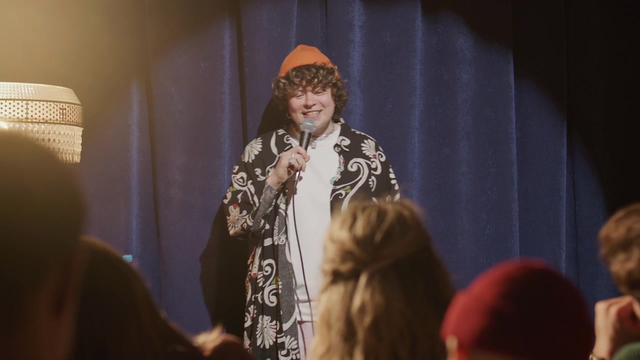 Kvindelig standupkomiker går ind på scenen og hilser på det klappende publikum
