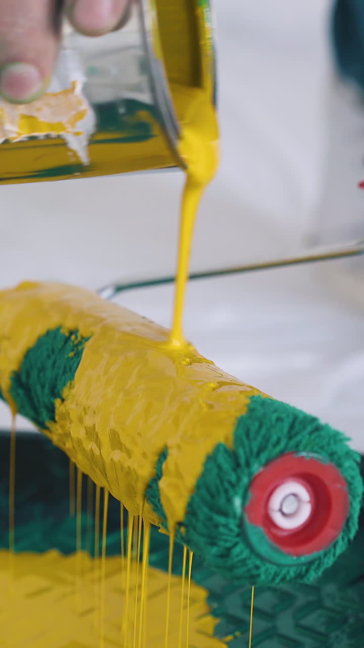 CU: professional builder pours bright yellow paint onto green roller above plastic tray on floor covered with foil closeup