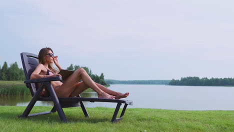 hermosa morena en traje de baño acostada en una tumbona en el lago sonriendo trabajando remotamente a través de internet en la esfera de los negocios. el concepto de la vida en el campo