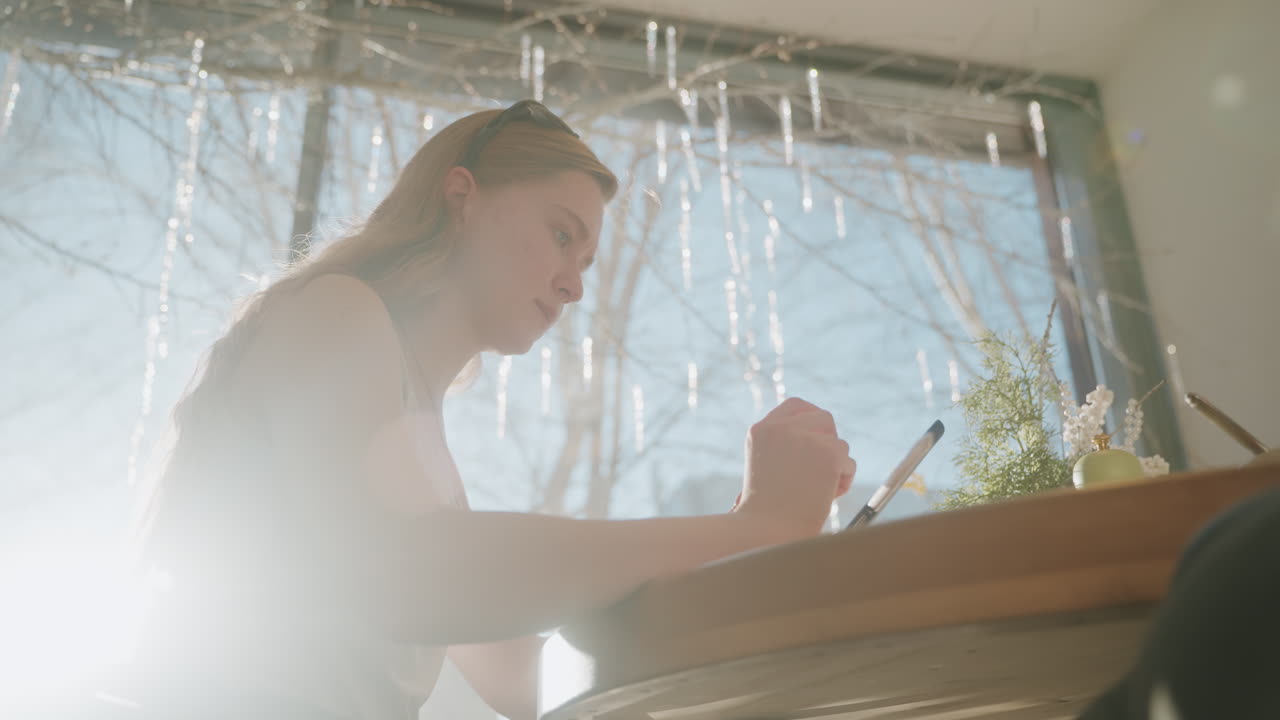 Solbeskinnet caféscene med kaukasisk kvinde, der skitserer på tablet med stylus, fokuseret profil ved stort vindue, nøgne grene synlige udenfor, varmt morgenlys og linseflare, lille plante og kaffe på bordet.