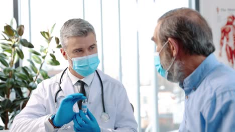 portrait of old pensioner male patient in medical mask sitting in hospital cabinet listening to middle aged experienced professional doctor showing and discussing vaccines on consultation 1