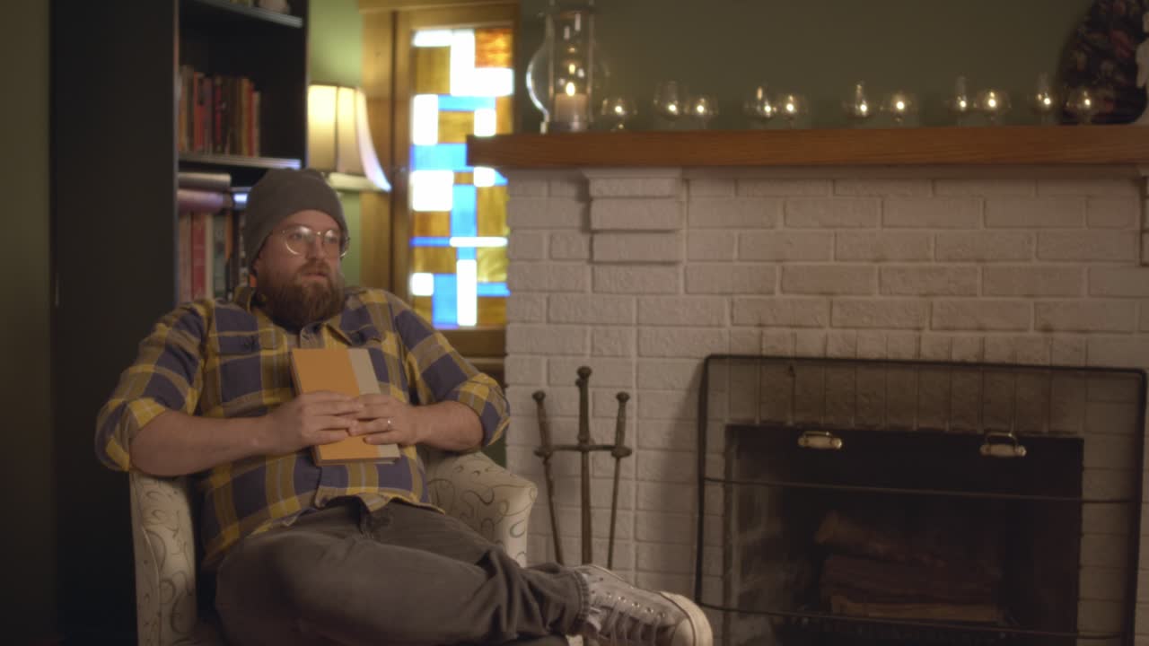 A man in plaid sitting, resting, and reading by the fireplace in a cozy fall living room setting, reading a book. Shot in 60 frames for slow motion