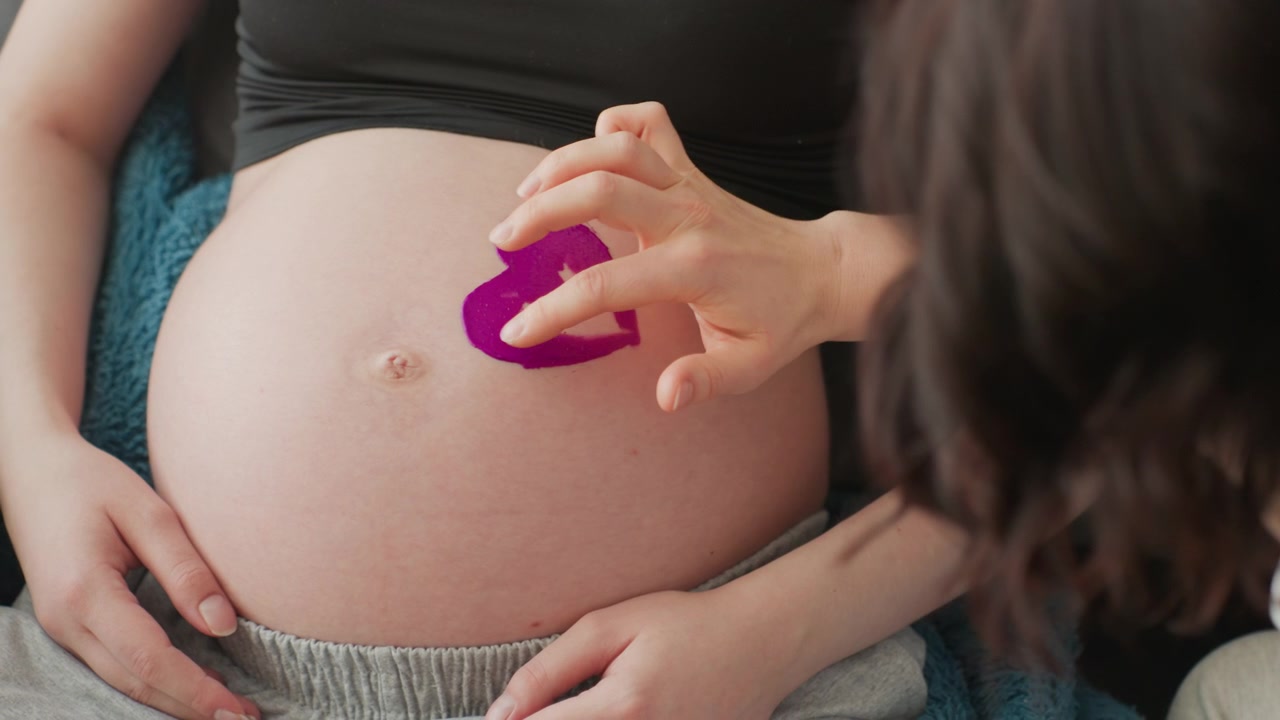 Primo piano di un bambino che disegna un cuore viola sul pancione di una donna incinta