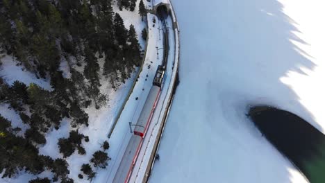 Antenne:-Blauer-Zahnradzug-In-Den-Pyrenäen,-Der-An-Einem-Zugefrorenen-See-Vorbeifährt-Und-In-Einen-Tunnel-Einfährt