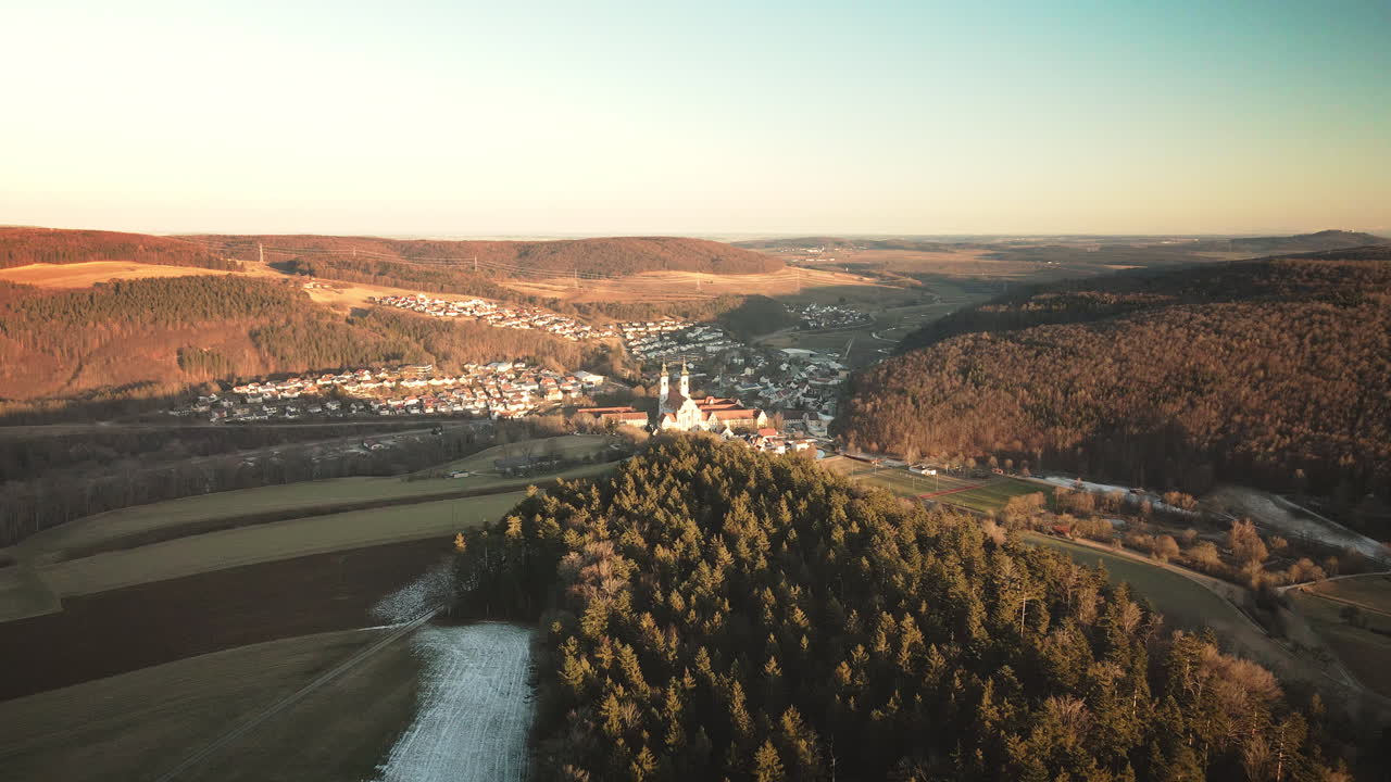 Drone glides forward toward Zwiefalten Abbey's twin towers on forested hill above town and winding river valley in autumnal Swabian Alb glow. Majestic baroque reveal