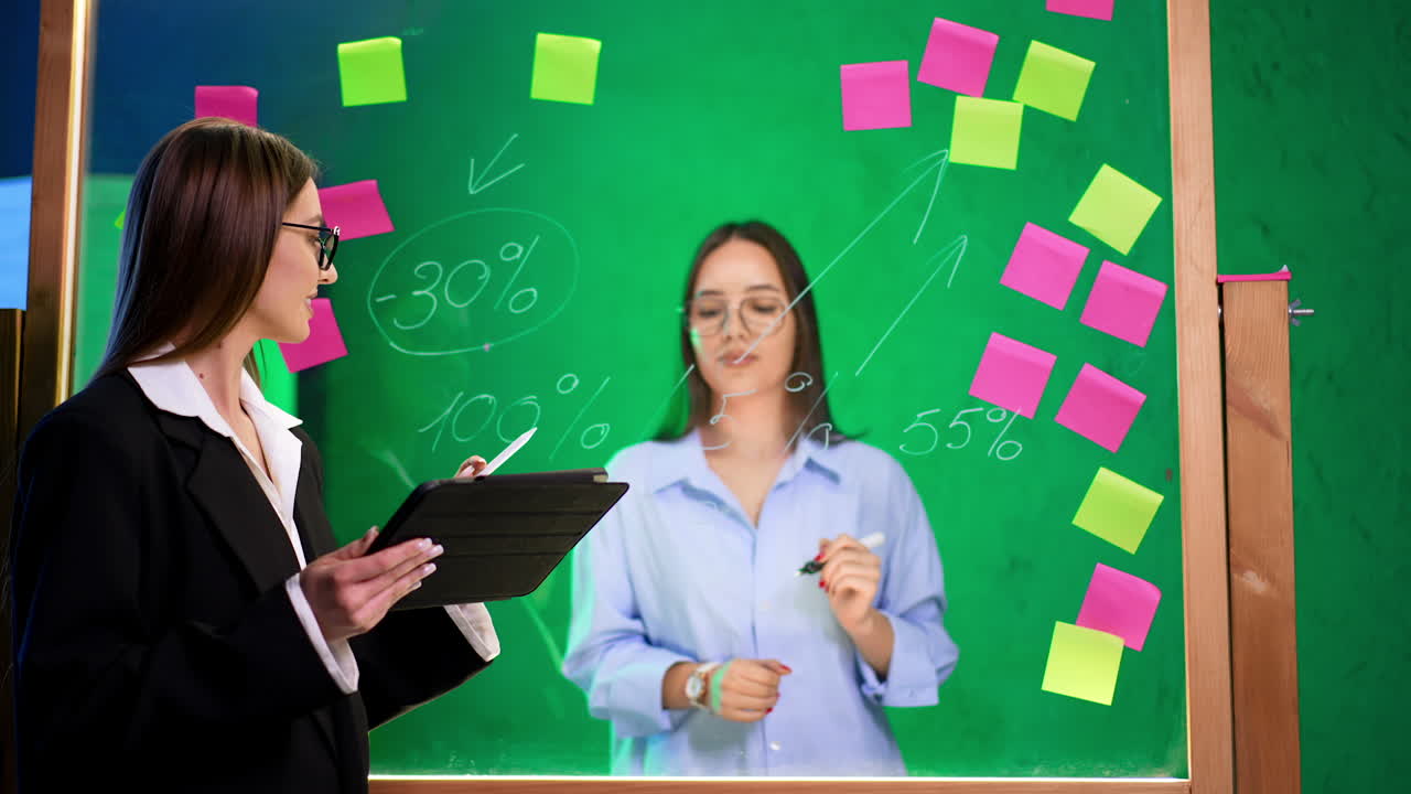 Brunette business lady wearing black jacket and holding a tablet stands at the glass board. Woman in white shirt writes numbers on the board.