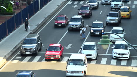 cars stopping at red light in down town main avenue of guayaquil (malecon) and then moving forward. some people passing by on sidewalk. a boy selling water bottles walks in the middle of the avenue.