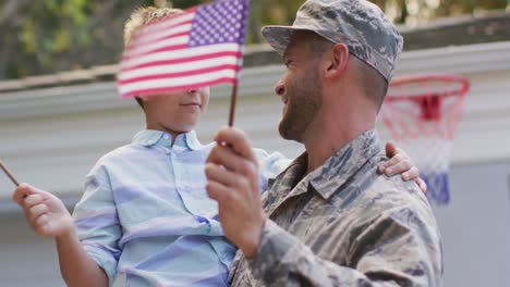 happy caucasian male soldier carrying his smiling son holding flag in garden outside their house