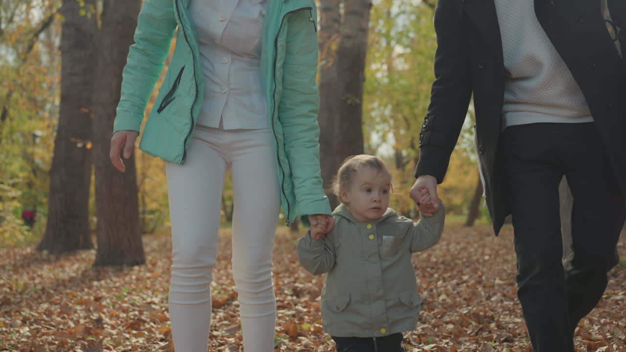 In un parco autunnale, genitori bianchi insegnano a un bambino a camminare, madre e padre tengono le mani del bambino, foglie scricchiolanti sotto i piedi, morbida luce dorata, giacche accoglienti e volti sorridenti, intimo momento familiare di celebrazione