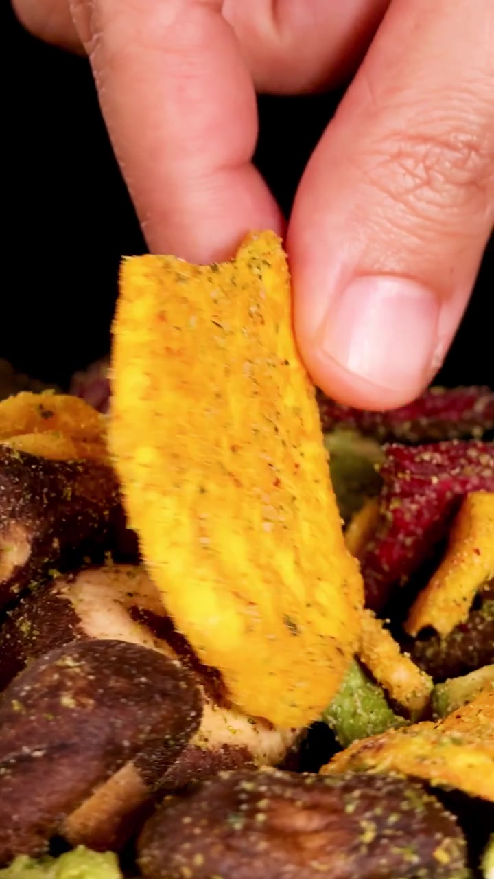 Uma mão alcança uma pilha colorida de chips de vegetais secos variados, pegando um. Iluminação de estúdio brilhante, fundo preto, close-up macro, movimento de câmera suave.