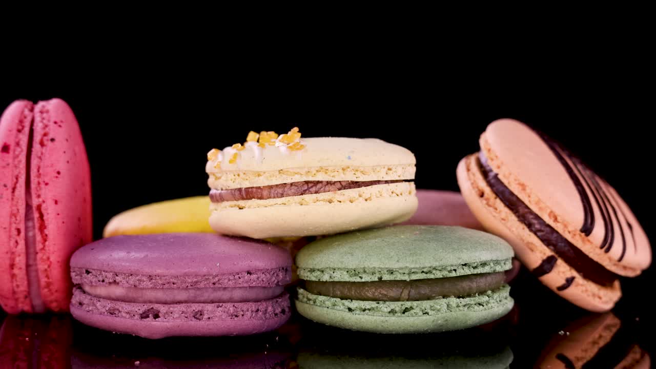 A hand reaches in to select a striped macaron from a vibrant assortment of French macarons on a glossy black surface under studio lighting