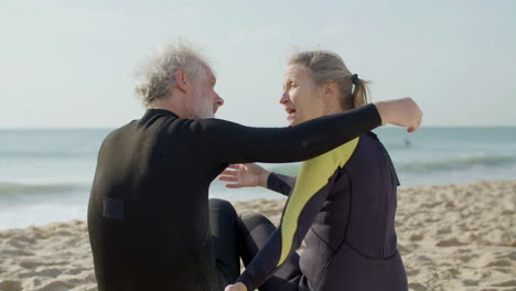 back view of a senior couple in wetsuit embracing while sitting on a surfboard at the beach 1