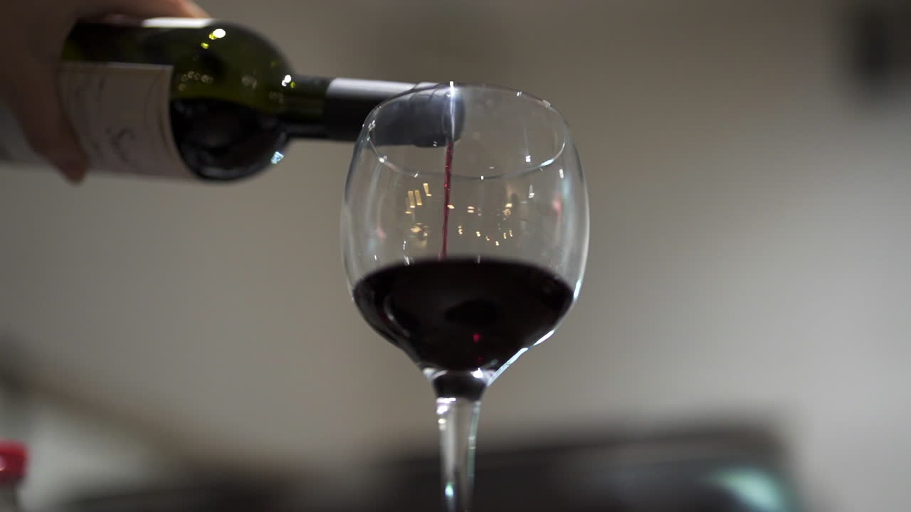 Detailed close-up shows red wine being poured from a bottle into a glass with soft focus on the flowing liquid and ambient indoor lighting, rich color and reflection, real time, slow motion shot