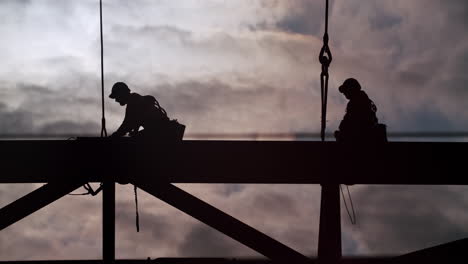 silhouettes of construction workers on a beam against a cloudy sky