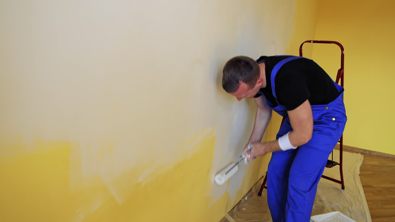 Young man paints a white wall with a roller. Repair of the interior. Male decorator painting a wall in the empty room. Painter in overalls doing renovation of the room.