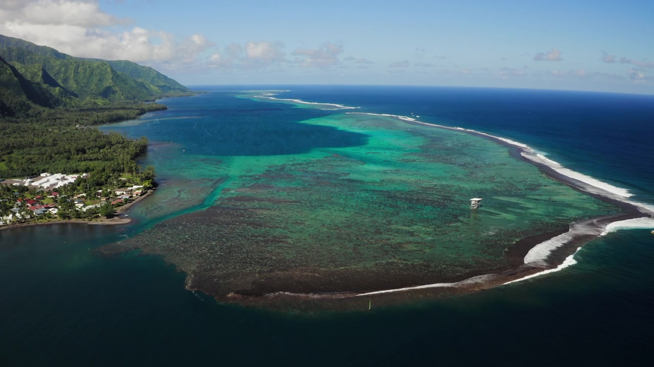 Drone che sorvola l'acqua bianca che si infrange sulla barriera corallina di Teahupoo, Tahiti, con il cielo azzurro chiaro che si riflette sulla laguna turchese circostante.
