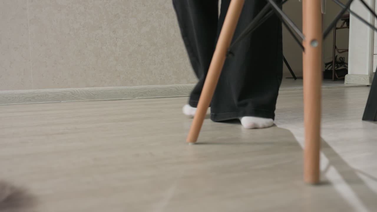 low angle view of woman carrying handbag as she approaches chair to drop bag while friend walks away from doorway across wood floor, capturing casual interior movement and visit moment