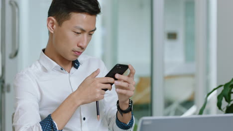 young asian businessman using smartphone texting checking messages on mobile phone sending email communication browsing online at work in office