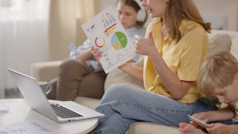 Female-Teacher-Having-Online-Class-At-Home-And-Showing-Charts-And-Statistics-While-Her-Children-Sitting-On-Sofa-Next-Her