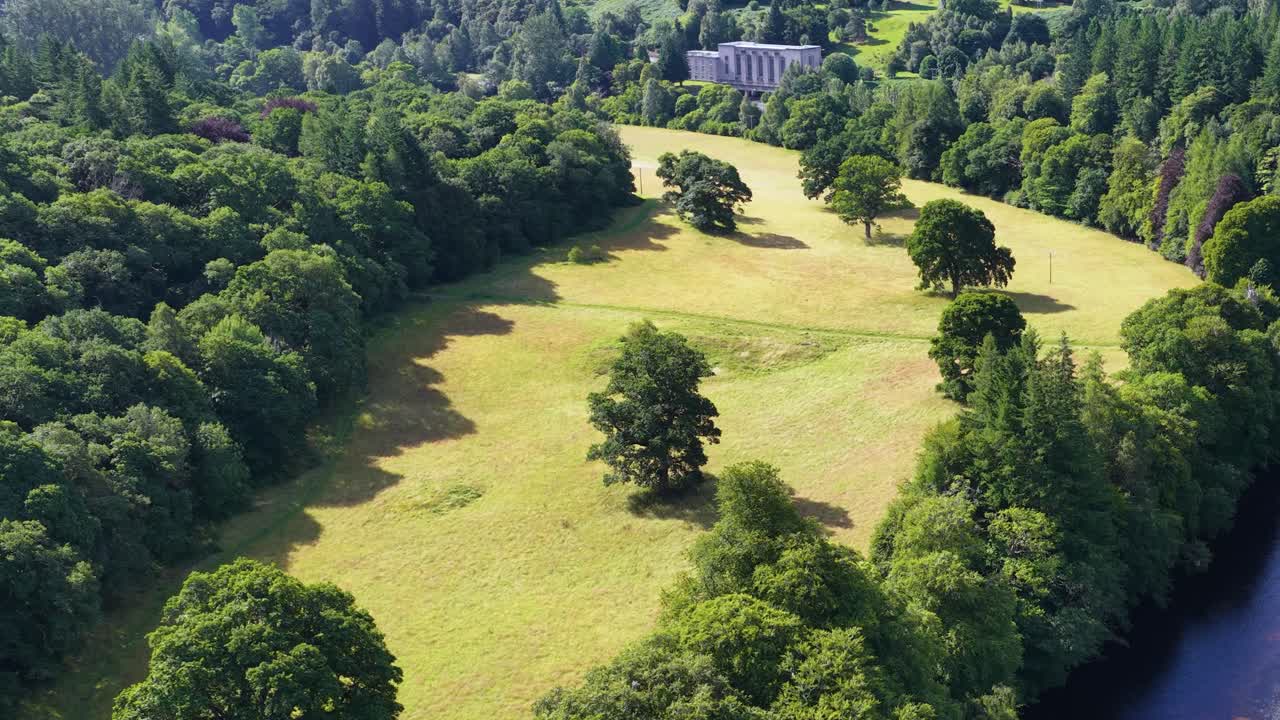 Drone glides above lush riverbank, revealing hydroelectric power station amid green Highlands landscape