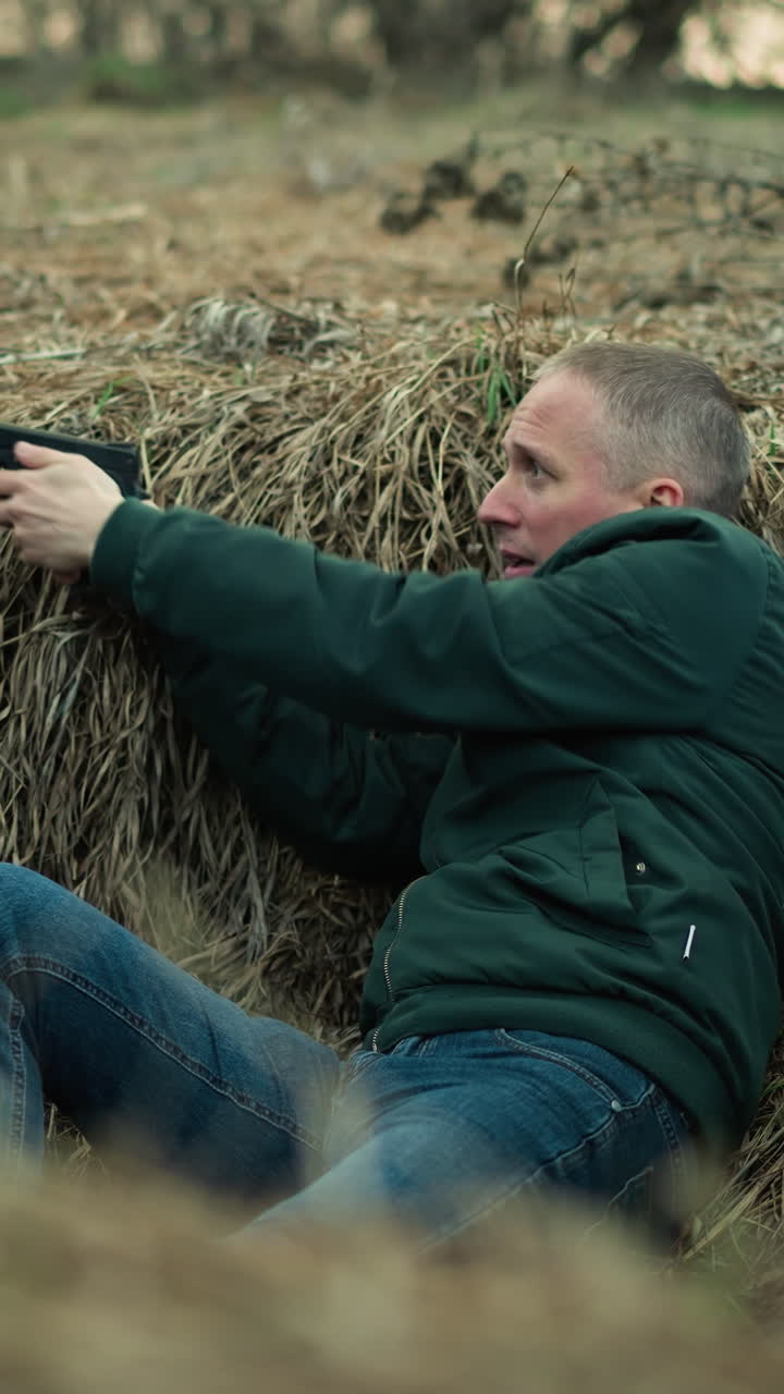 una vista de cerca de un hombre con una chaqueta verde y vaqueros sosteniendo una pistola mientras yacía en una zanja seca y cubierta de hierba, mirando tenso asustado