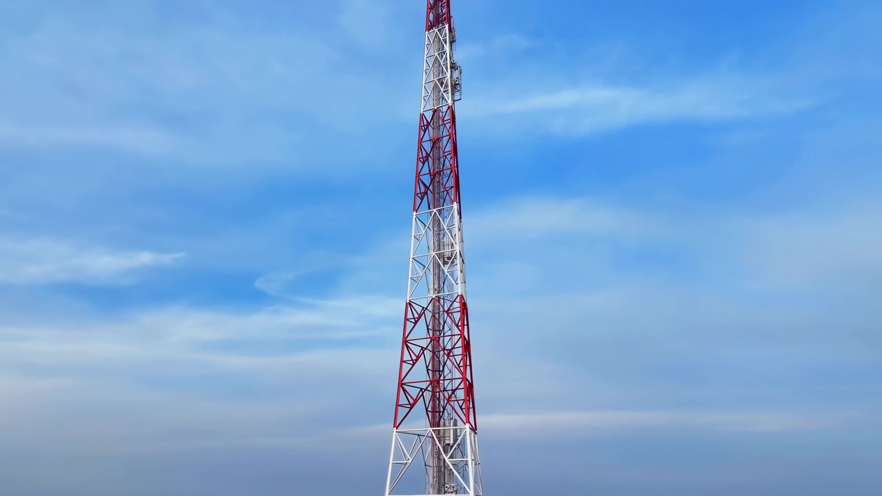 Sakin kırsal tarlaların üzerinde yükselen uzun kırmızı beyaz kafes iletişim kulesi, ince bulutlu berrak mavi gökyüzünün altında, açık ufuk ve geniş kırsal manzarayla birlikte. Kule, anten, yapı.