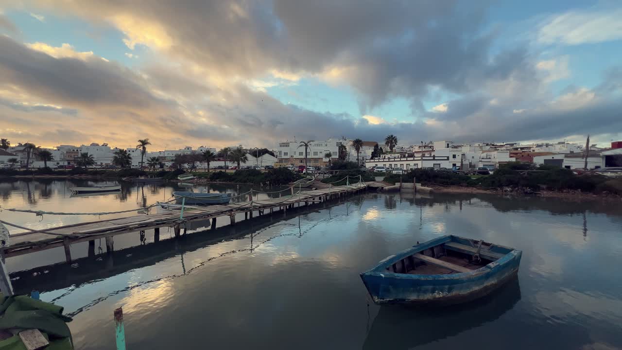la desembocadura del río barbate en barbate, andalucía, españa, refleja el cielo nublado al atardecer, con viejos barcos de pesca de madera y un muelle creando una escena pacífica.