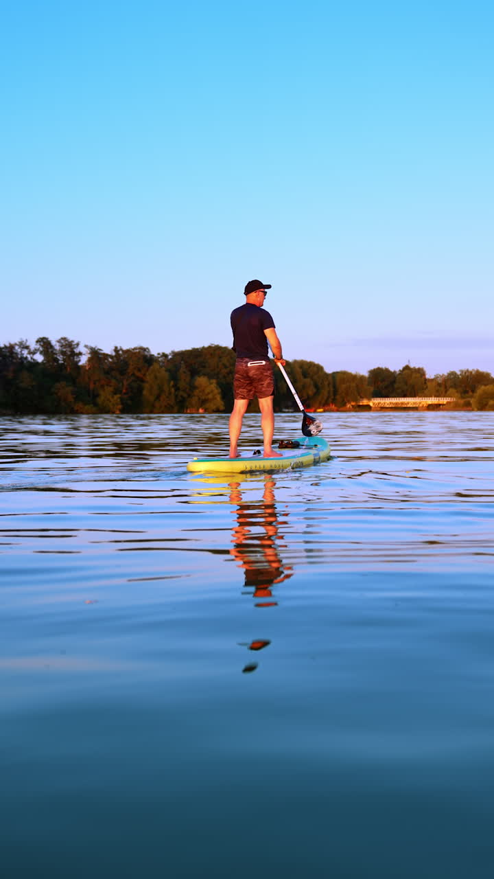 Rear view on the man rowing with an oar on sup board. Low angle view on the sportsman having active time spending in the nature at sunset. Low angle view. Vertical video.