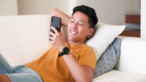 Young man relaxing on couch using smartphone, looking thoughtful at home