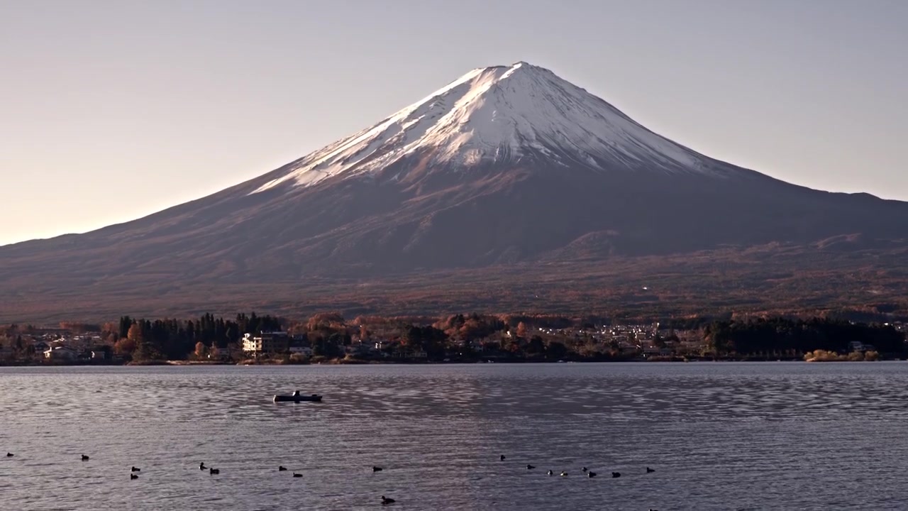 Una toma de inclinación ascendente suave y cinematográfica que comienza desde botes de remos tradicionales flotando en el lago Kawaguchiko, ascendiendo sobre el agua para revelar el Monte Fuji con su pico nevado en Japón.