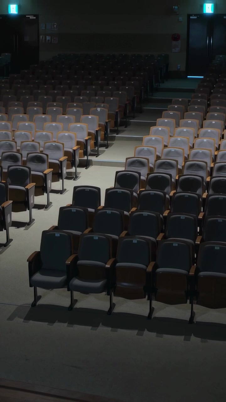 Video verticale: Telecamera che si inclina rivelando sedie imbottite in un auditorium poco illuminato per mostrare le uscite di sicurezza.