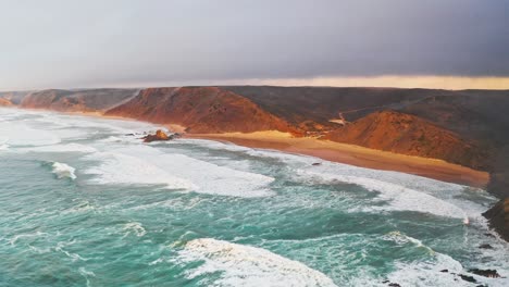 Portugal beach cliffs on Atlantic ocean at sunset from high aerial drone pull back shot with rough waves