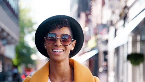 millennial black woman wearing a yellow pea coat, sunglasses and a homburg hat, standing on a city street smiling to camera, close up