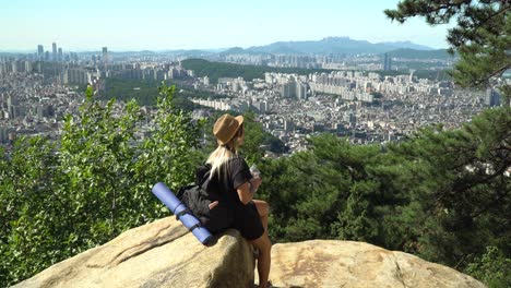 niña sentada en la roca y bebiendo después de una larga caminata y mirando hacia el horizonte del centro del distrito de seocho-gu desde la montaña gwanaksan en seúl, corea del sur